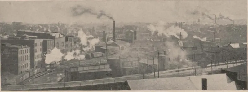 A wide view over rooftops of a crowded factory district, with brick mill buildings, rail yards and tall smokestacks sending up plumes of smoke and steam.