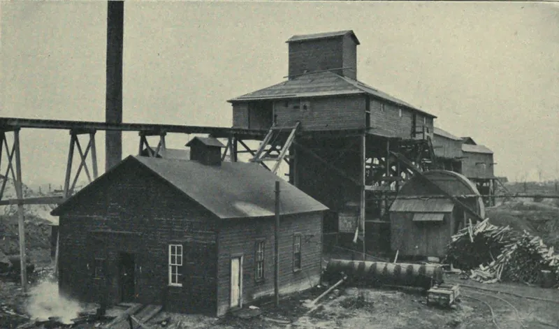 A wooden engine house in front of a tall wooden tipple and shaft house, with a trestle running off to the left, a smokestack, a drum-shaped fan housing at right and a stack of mine timbers.