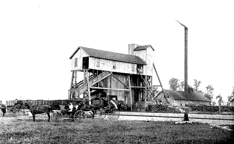 A tall wooden coal tipple raised on timber legs, with a tall smokestack and a long low shed behind it, horse-drawn buggies in front and loaded coal cars on a siding.