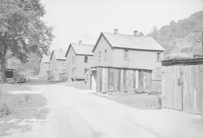 A dirt road running past a row of identical two-story wooden company houses at the foot of a wooded hill, with a wooden shed and fence in the foreground and a parked car in the distance.