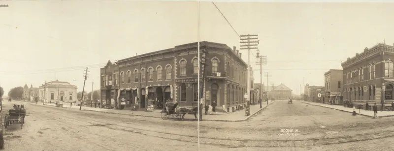 A wide panoramic view of a street intersection with brick bank and business buildings, a frame storefront at left, utility poles, streetcar tracks in the unpaved street and a horse-drawn buggy.