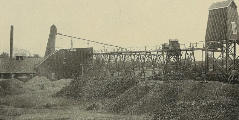 A long wooden trestle tramway runs from a tall shaft house on the right to a barn-like mill building marked The Monaco, with heaps of crushed rock in the foreground and a smokestack at left.