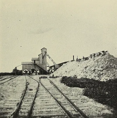 A wooden coal tipple with an inclined trestle rises beside a tall bank of mine waste, with several railroad tracks curving toward it in the foreground.