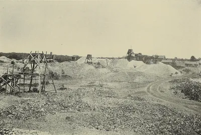 A rough field of broken rock and pale waste heaps, with a small wooden hand windlass derrick in the foreground and more derricks, sheds and trees in the distance.