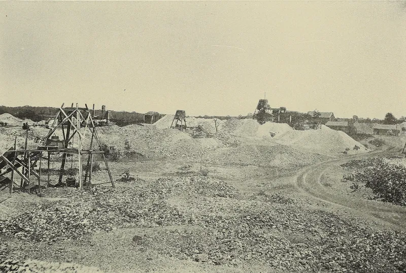 A rough field of broken rock and pale waste heaps, with a small wooden hand windlass derrick in the foreground and more derricks, sheds and trees in the distance.