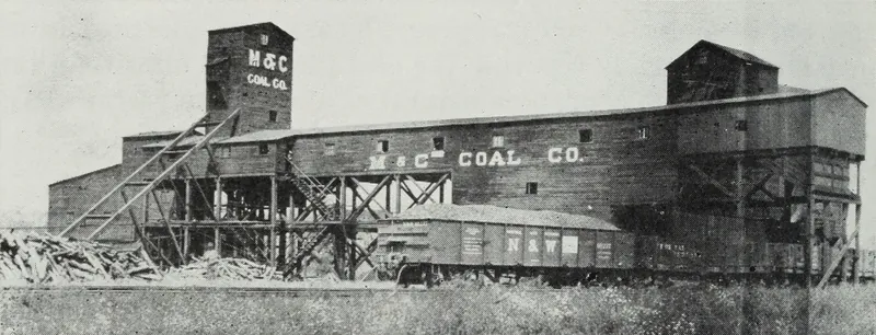 A long wooden tipple and screening building painted with the words "M & C Coal Co.", with a tall shaft tower at left, timber bracing, piled lumber, and railroad coal cars beneath.