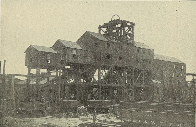 A massive wooden tipple and screening house on timber legs, topped by a headframe wheel, with railroad coal cars and a few workmen below it.