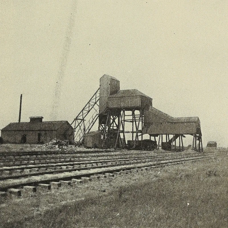 A small wooden tipple with an inclined trestle and a low brick engine house beside several railroad sidings on open prairie.