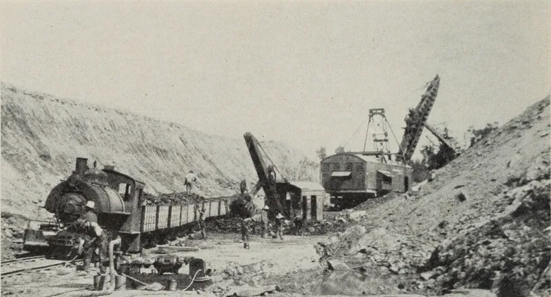A coal strip pit between high banks of earth, with a small steam locomotive pulling coal cars at left and two large electric shovels working at right, men standing between them.