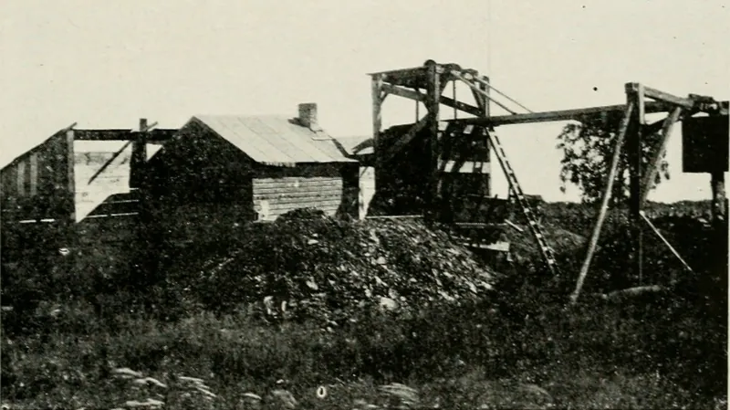 A small coal mine surface plant: a wooden headframe with ladders and a hoisting beam beside a shed with a brick chimney, behind a heap of coal and weeds.