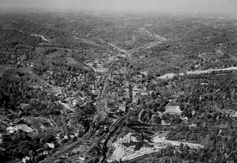 An aerial photograph of a small town in a narrow valley among wooded hills, with rail lines running through the center and a large pale waste pile beside them in the foreground.