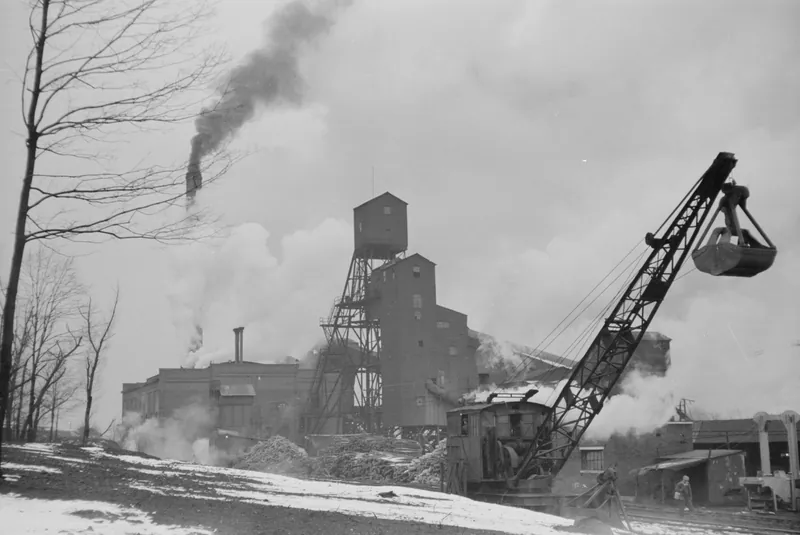A brick mine building and steel headframe wreathed in steam and smoke, with a crane and clamshell bucket at right and snow on the ground.