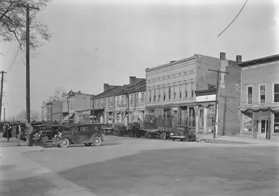 A wide dirt main street lined with two- and three-story brick and frame storefronts, with parked cars, trucks and a few people along the sidewalk.