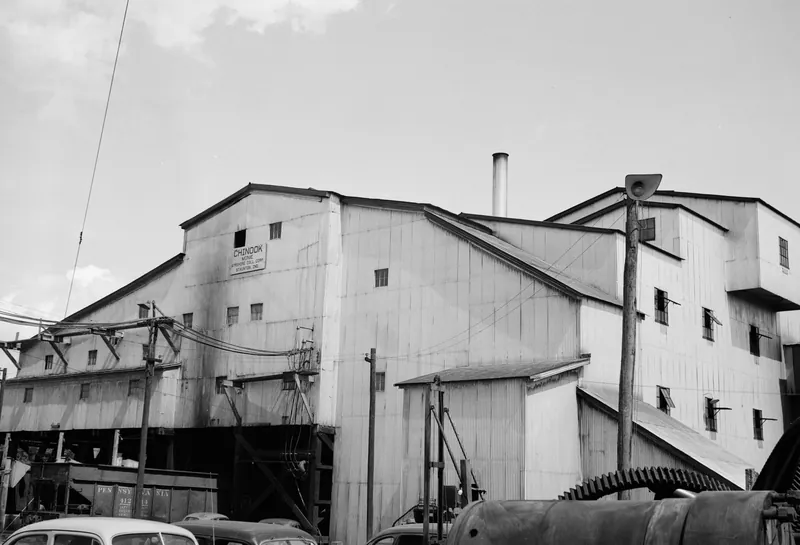 A large sheet-metal coal tipple building with many small windows, power poles and wires in front, a big gear and machinery in the foreground, and parked cars at the bottom.