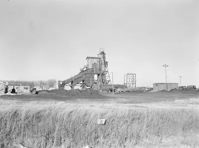 A wooden coal mine headframe and tipple with a long conveyor, coal piles and parked cars, seen across a field of tall grass.