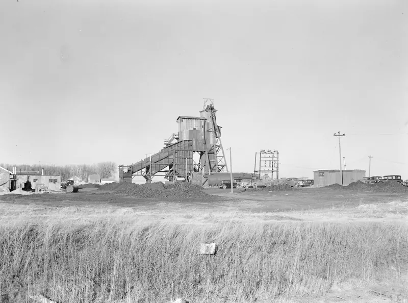 A wooden coal mine headframe and tipple with a long conveyor, coal piles and parked cars, seen across a field of tall grass.