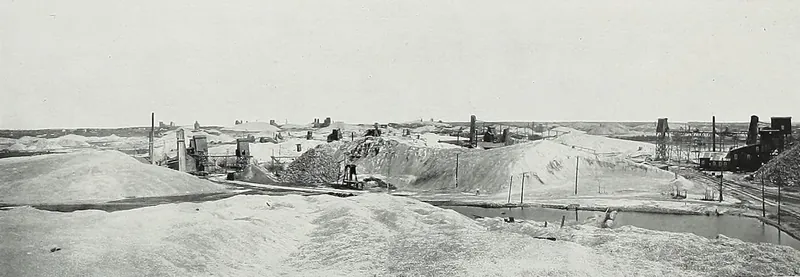 A wide view across pale mounds of crushed rock waste, with wooden mine derricks, mill buildings, smokestacks and a pond of water spread over flat ground.