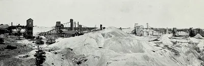 Wooden shaft houses, ore bins, mill buildings and smokestacks stand along a hillside of pale mine waste, with a small evergreen tree in the foreground.