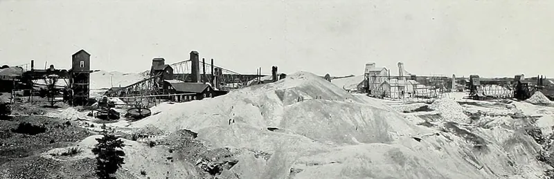 Wooden shaft houses, ore bins, mill buildings and smokestacks stand along a hillside of pale mine waste, with a small evergreen tree in the foreground.