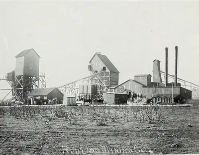 Tall wooden derrick houses, an inclined conveyor and long mill buildings with two smokestacks, seen across a scrubby field, with horses and wagons near the buildings. The words Red Dog Mining Co. are written on the photograph.