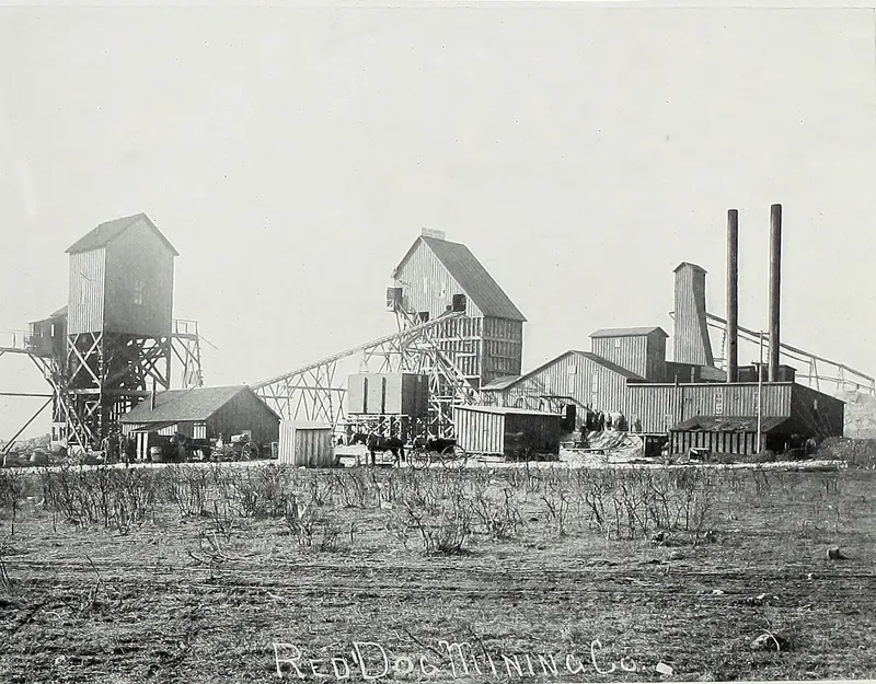 Tall wooden derrick houses, an inclined conveyor and long mill buildings with two smokestacks, seen across a scrubby field, with horses and wagons near the buildings. The words Red Dog Mining Co. are written on the photograph.