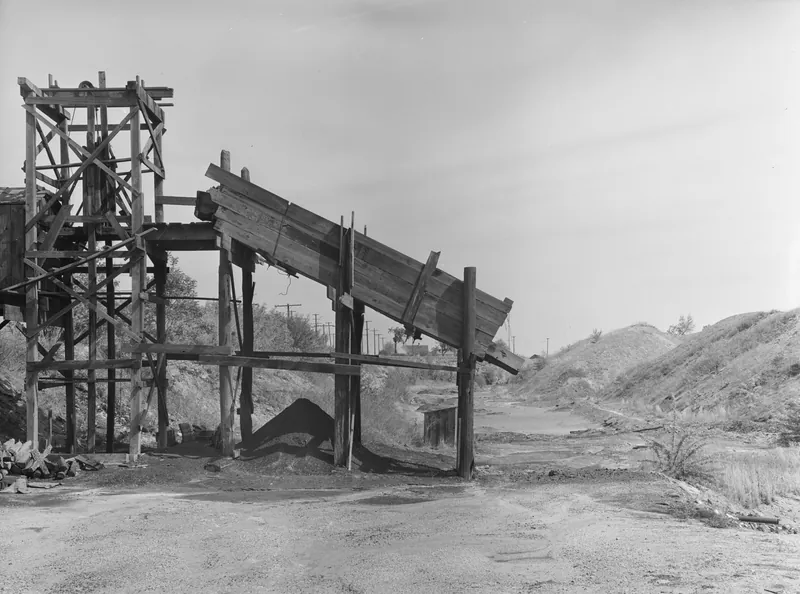 A weathered wooden mine headframe and an inclined coal chute beside a dirt road, with large mine waste dumps on the right.