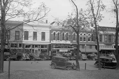A row of two-story brick and stone storefronts, including a theater marked Temple, with 1930s cars parked in front and bare trees in the foreground.