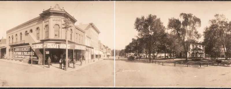 A wide panoramic view of a small-town square with a park and bandstand under large trees, a corner clothing store, two-story brick storefronts, horses and buggies, and men standing on the walks.
