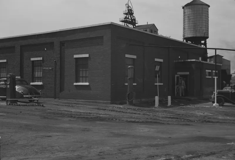 A one-story brick mine washhouse on a dirt lot, with a steel mine headframe and a wooden water tank rising behind it, a gas pump, and a man standing in the doorway.