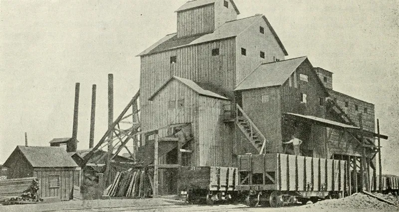 A tall wooden coal tipple and screening house with stairways, a small shed at left, three smokestacks, and loaded railroad coal cars along the front.