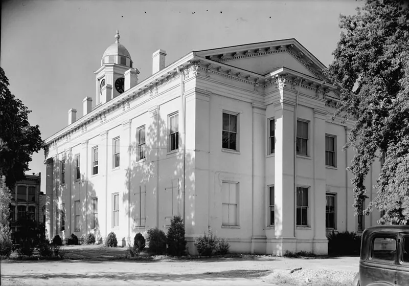 A large white two-story courthouse with tall pilasters, a pediment and a domed clock cupola, shaded by trees, with a 1930s automobile parked at lower right.