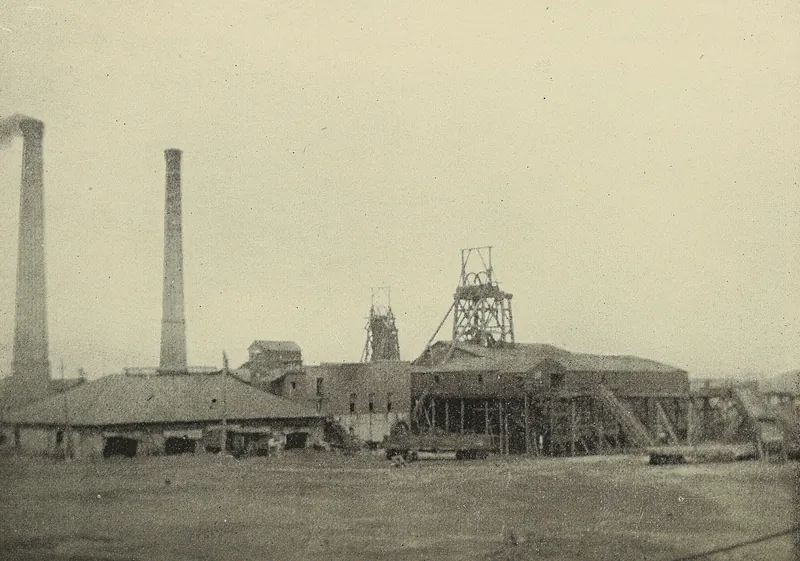 Two tall brick smokestacks rise beside low mine buildings, with a steel headframe and a wooden one above the shaft house and an open yard in front.