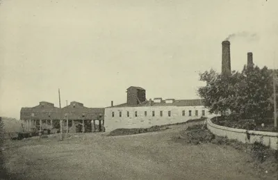 A long two-story stone building and dark wooden mine structures beside a dirt road, with two smokestacks and a grove of trees at right and a curving stone wall.