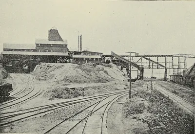 Railroad tracks fan out across a yard toward a mine plant with a tall shaft house, long sheds, mounds of coal and an elevated trestle with coal cars.