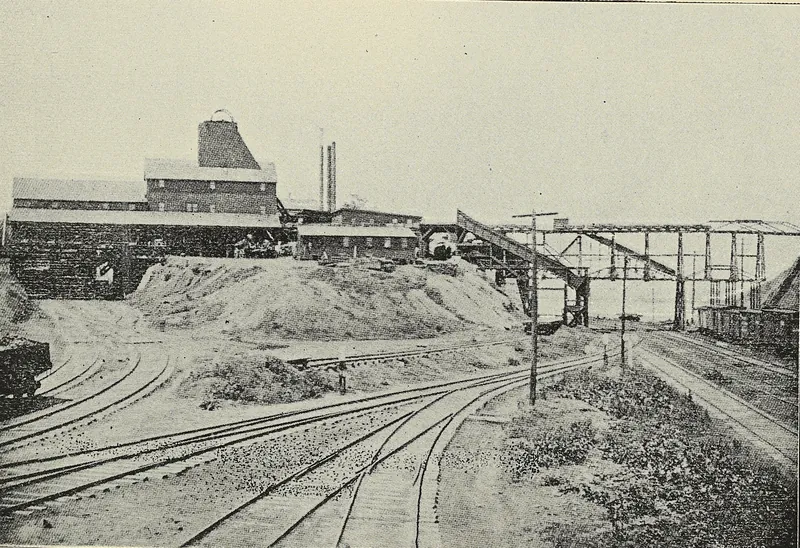 Railroad tracks fan out across a yard toward a mine plant with a tall shaft house, long sheds, mounds of coal and an elevated trestle with coal cars.