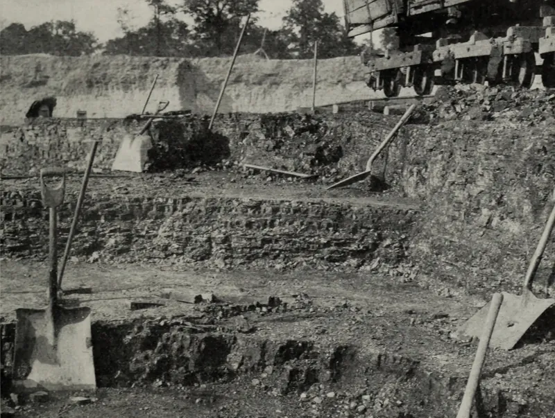 An open clay and coal pit with stepped benches of dark coal, shovels and a pick leaning on the ledges, and a line of side-dump cars on a track at upper right.