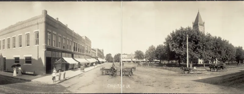 A wide panoramic view of a town square: brick business blocks on two sides, a courthouse tower behind tall trees, unpaved streets, horse-drawn wagons and people on the sidewalks.
