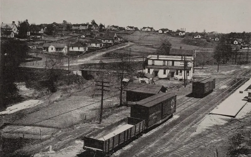 A view from above of a small mining town: railroad tracks with boxcars and an open coal car in front, a two-story frame building, a road climbing a hill, and rows of frame houses along the ridge.