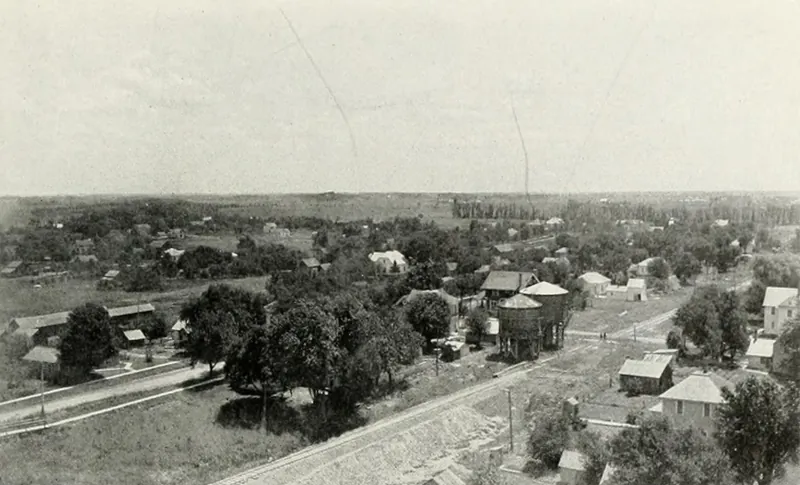 A view over treetops and houses of a small prairie town, with a railroad embankment curving across the foreground and flat farmland to the horizon.