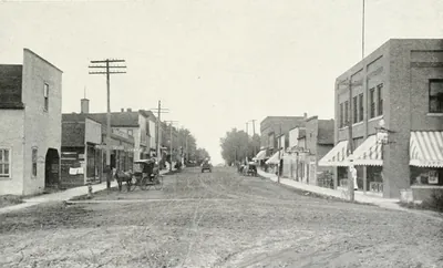 An unpaved main street lined with brick and frame storefronts with striped awnings, a horse and buggy, and telephone poles.