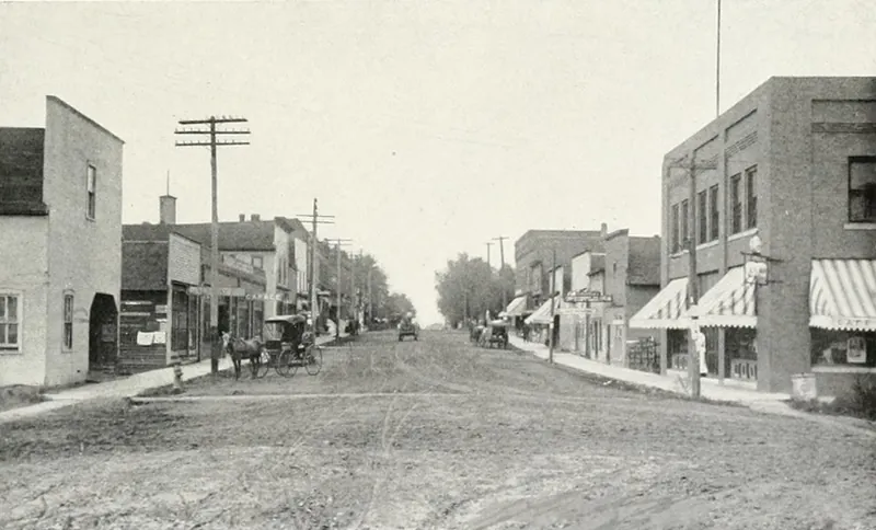 An unpaved main street lined with brick and frame storefronts with striped awnings, a horse and buggy, and telephone poles.