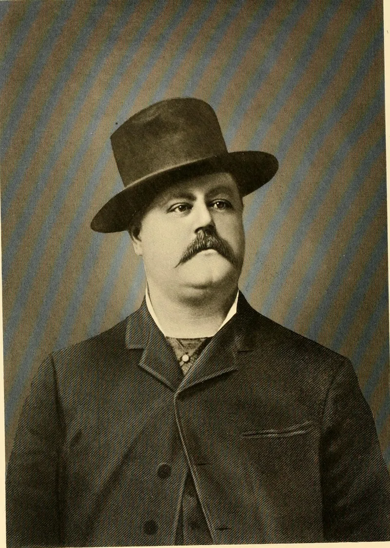 Studio portrait of a heavy-set man with a large mustache, wearing a tall dark hat, a dark coat buttoned high and a patterned necktie.
