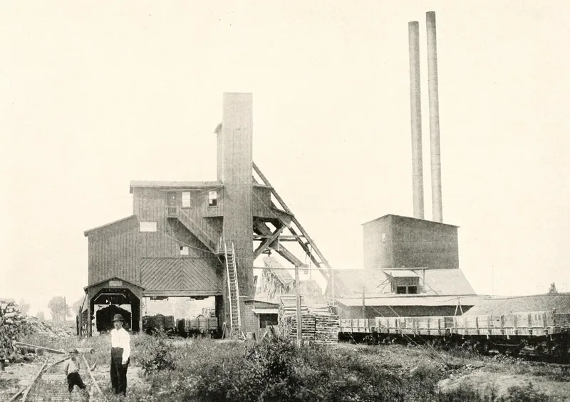 A tall wooden coal mine shaft house with an inclined trestle, two tall smokestacks behind a boiler house, a line of railroad cars, and a man and a boy standing in the weeds in the foreground.