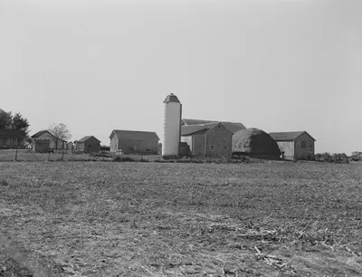 A farmstead across a harvested field, with a tall silo, several barns and sheds, and a large haystack.