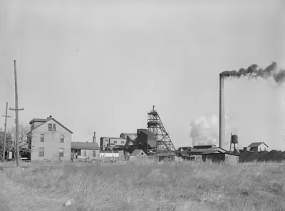 Frame houses in the foreground and a steel mine headframe, mine buildings and a smoking stack behind, seen across a grassy field.