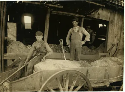 A boy in a cap sits on a wagon full of crushed ore holding a long tool, while a man in overalls stands with a shovel inside an open mine shed.