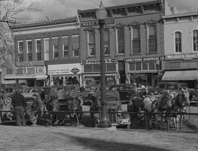 Horse teams and farm wagons tied up beside parked automobiles at a street lamp in front of a row of brick storefronts, including Armstrong Hardware and a market.