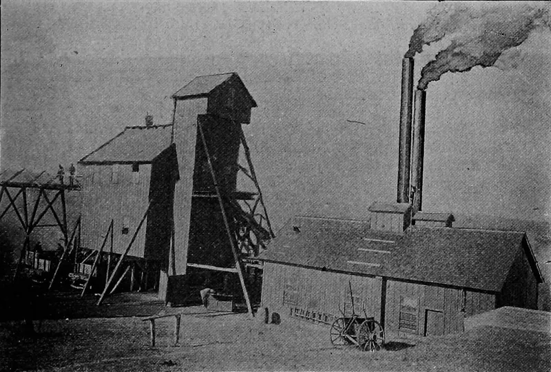 A tall wooden shaft house and tipple beside a long, low engine house with two tall smokestacks pouring smoke; a farm wagon stands in front.