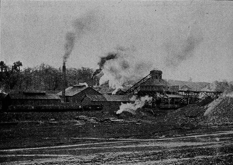 Low mine buildings and a tipple across open, muddy ground, with a tall smokestack and several plumes of smoke and steam.