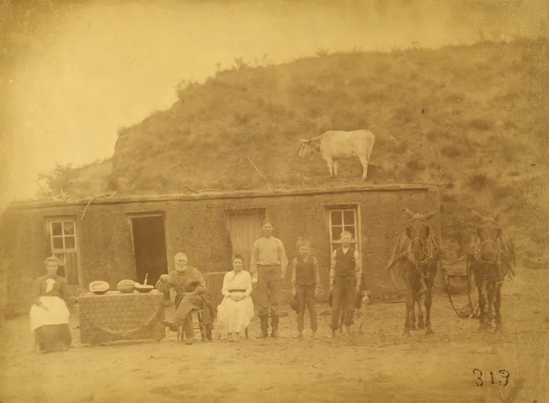 A family seated and standing in front of a sod house dug into a hillside, with a cow standing on the sod roof, a table of melons, and a team of mules at right.
